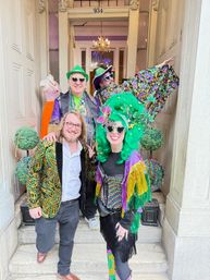 Four people in colorful Mardi Gras-style costumes and beads posing on the steps of a historic storefront entrance, featuring a large green wig, sequined jackets, masks, fringe and decorative topiary balls