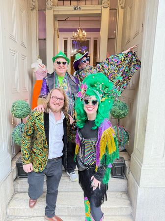 Four people in colorful Mardi Gras-style costumes and beads posing on the steps of a historic storefront entrance, featuring a large green wig, sequined jackets, masks, fringe and decorative topiary balls