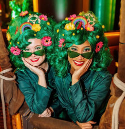 Two smiling costumed performers in bright emerald wigs decorated with flowers, rainbows and gears, wearing green sunglasses and teal jackets, leaning on a wicker hot-air-balloon basket with ropes.
