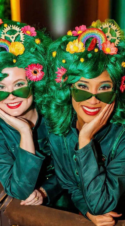 Two smiling costumed performers in bright emerald wigs decorated with flowers, rainbows and gears, wearing green sunglasses and teal jackets, leaning on a wicker hot-air-balloon basket with ropes.