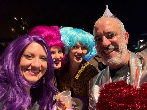 Nighttime group selfie at a costume party: three women in purple, magenta and turquoise wigs and a man with a glittery beard and tiny silver cone hat, all smiling and holding a plastic cup.