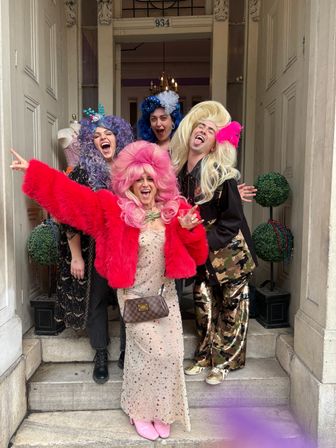 Four exuberant partygoers in colorful wigs and sparkly costumes laughing and posing in a building doorway flanked by decorative topiary balls — playful, festive group photo