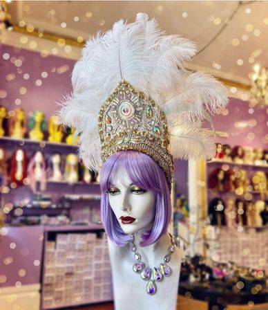 Glam mannequin with a purple bob wig wearing an ornate gold and crystal costume headdress topped with white feathers and an iridescent teardrop necklace on a boutique display