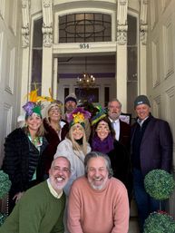 Group of smiling people in a decorative doorway wearing colorful feathered headpieces and glitter face jewels in purple, green and gold, with stained-glass transom and chandelier visible inside.