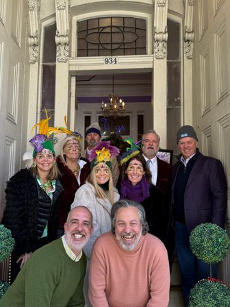 Group of smiling people in a decorative doorway wearing colorful feathered headpieces and glitter face jewels in purple, green and gold, with stained-glass transom and chandelier visible inside.