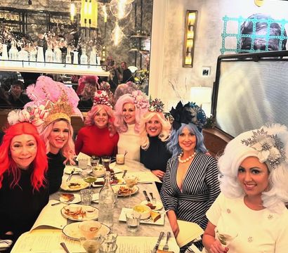 Group of women in colorful wigs and elaborate headpieces enjoying brunch at a marble-topped table in a stylish bistro, with cocktails, plates of food and a mirrored wall reflecting the cozy, decorative interior.