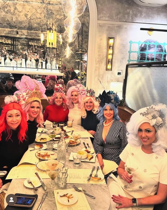 Group of women in colorful wigs and elaborate headpieces enjoying brunch at a marble-topped table in a stylish bistro, with cocktails, plates of food and a mirrored wall reflecting the cozy, decorative interior.