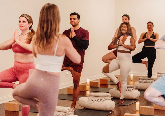 Diverse group yoga class in a cozy indoor studio practicing tree pose on mats with bolsters, candles and blankets — smiling participants balancing in a guided session