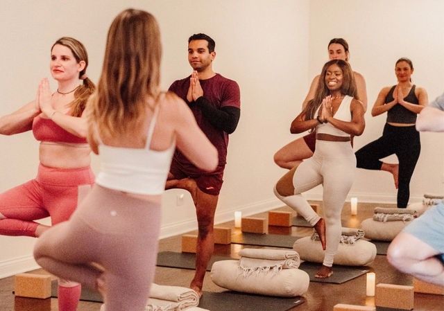 Diverse group yoga class in a cozy indoor studio practicing tree pose on mats with bolsters, candles and blankets — smiling participants balancing in a guided session
