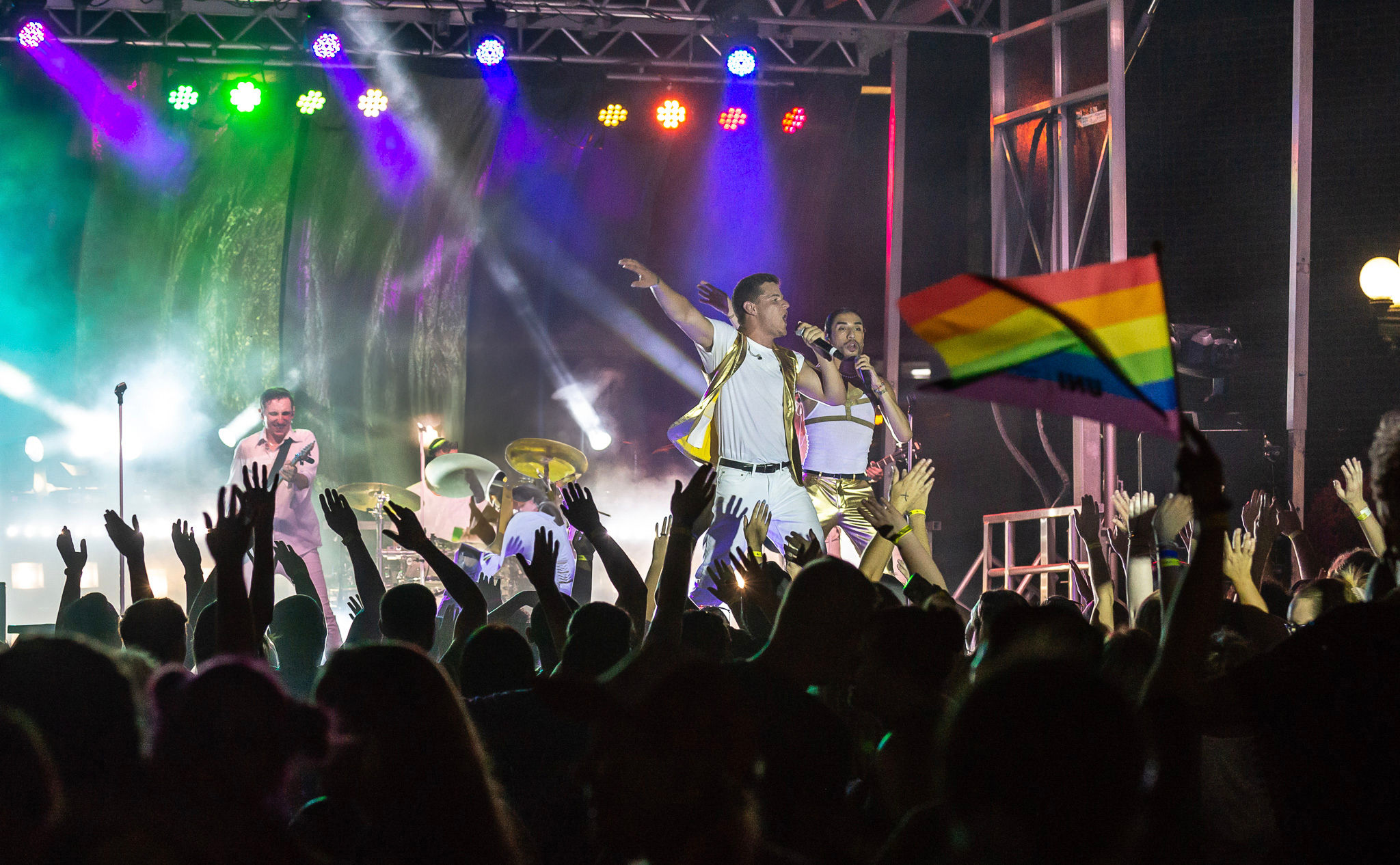 Nighttime outdoor concert with a cheering crowd and raised hands as a live band performs in white and gold on a colorful lit stage, a rainbow pride flag waving in the audience.