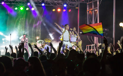 Nighttime outdoor concert with a cheering crowd and raised hands as a live band performs in white and gold on a colorful lit stage, a rainbow pride flag waving in the audience.