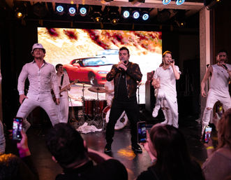 Energetic indoor pop concert with a lead singer in a black jacket, backup dancers in white, drummer and guitarist in front of a colorful LED backdrop, bright stage lights overhead and audience filming on smartphones.