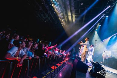 Excited crowd pressed at a barrier in an indoor live music venue, reaching toward a male vocal group in white suits performing on a brightly lit stage with colorful spotlights
