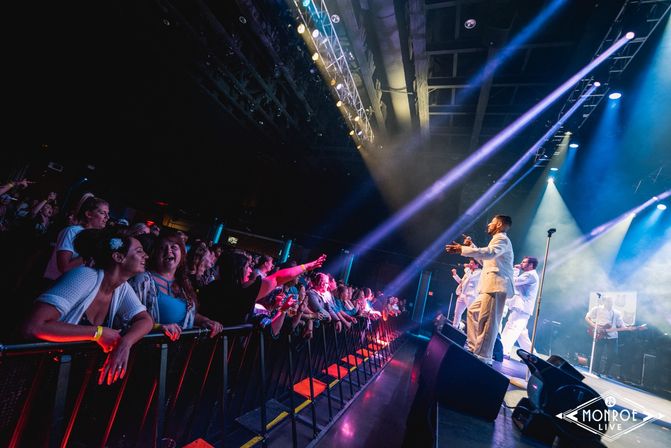 Excited crowd pressed at a barrier in an indoor live music venue, reaching toward a male vocal group in white suits performing on a brightly lit stage with colorful spotlights