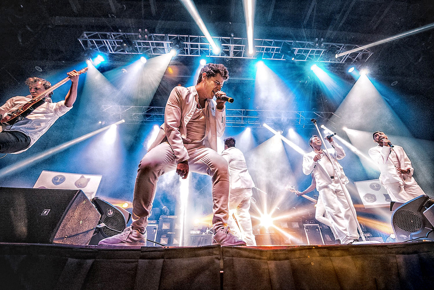 Low-angle shot of a high-energy pop band on an indoor concert stage — singers in white suits and a leaping guitarist performing amid colorful spotlights, smoke, and dramatic stage lighting.