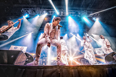 Low-angle shot of a high-energy pop band on an indoor concert stage — singers in white suits and a leaping guitarist performing amid colorful spotlights, smoke, and dramatic stage lighting.