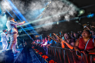 Live indoor concert with a singer in a white suit on a smoky, blue-lit stage while an energetic front-row crowd cheers, reaches forward and records on phones at a packed venue.