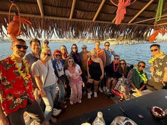 Group of friends wearing leis on a tiki-style party boat at a sunny marina, posing under a thatched roof with sailboats and harbor in golden-hour light.