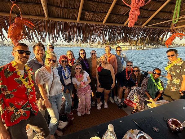 Group of friends wearing leis on a tiki-style party boat at a sunny marina, posing under a thatched roof with sailboats and harbor in golden-hour light.