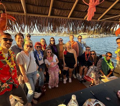 Group of friends wearing leis on a tiki-style party boat at a sunny marina, posing under a thatched roof with sailboats and harbor in golden-hour light.