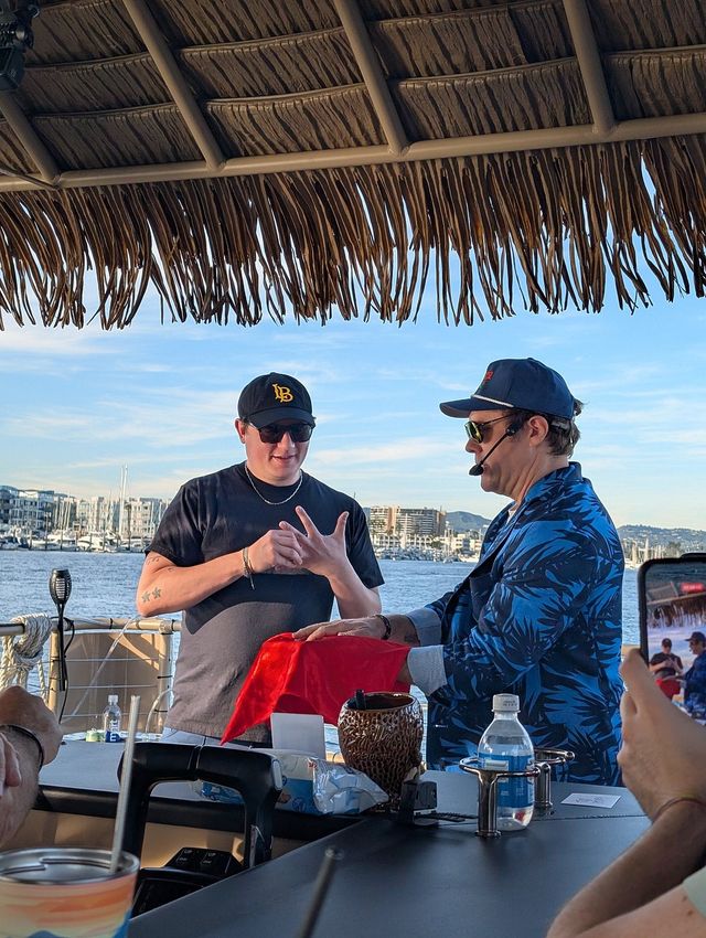 Performer in a blue tropical jacket and headset doing a red-cloth magic trick for a spectator in a black cap under a tiki-thatched boat canopy with a marina and skyline in the background.