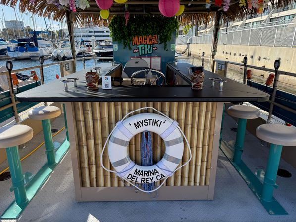 Floating tiki bar docked at a coastal marina — bamboo-front counter with life preserver, swivel bar stools, colorful lanterns, and moored boats