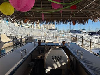 Thatched-roof tiki boat helm overlooking a sunny coastal marina with docked sailboats, colorful paper lanterns hanging from the roof, steering wheel and bar counters in the foreground