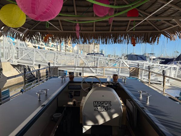 Thatched-roof tiki boat helm overlooking a sunny coastal marina with docked sailboats, colorful paper lanterns hanging from the roof, steering wheel and bar counters in the foreground