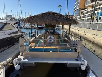 Floating tiki-bar boat with a thatched roof and bar stools docked among yachts at a sunny coastal marina with palm trees
