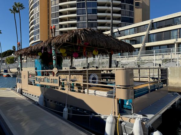 Tiki-themed party boat with a thatched roof and colorful lanterns docked at a sunny marina beside waterfront high-rise condos and palm trees