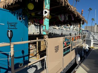 Tiki-themed floating bar moored at a sunny marina dock with a thatched roof, carved tiki posts, colorful lanterns, palm trees and nearby harbor buildings.