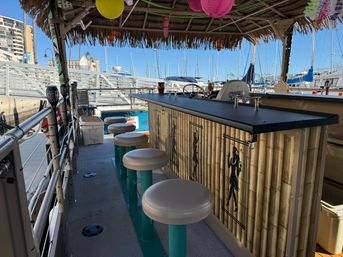 Tiki-style floating bar on a pontoon boat with bamboo façade, round teal stools and paper lanterns overhead, docked at a sunny marina with sailboat masts and waterfront buildings
