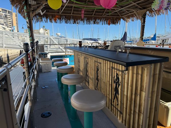 Tiki-style floating bar on a pontoon boat with bamboo façade, round teal stools and paper lanterns overhead, docked at a sunny marina with sailboat masts and waterfront buildings