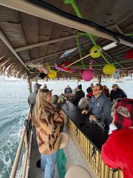 Passengers chatting at a tiki-style floating bar cruise in a coastal harbor — thatched roof decorated with colorful paper lanterns and leis, bamboo counter and ocean views.