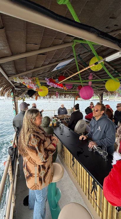 Passengers chatting at a tiki-style floating bar cruise in a coastal harbor — thatched roof decorated with colorful paper lanterns and leis, bamboo counter and ocean views.
