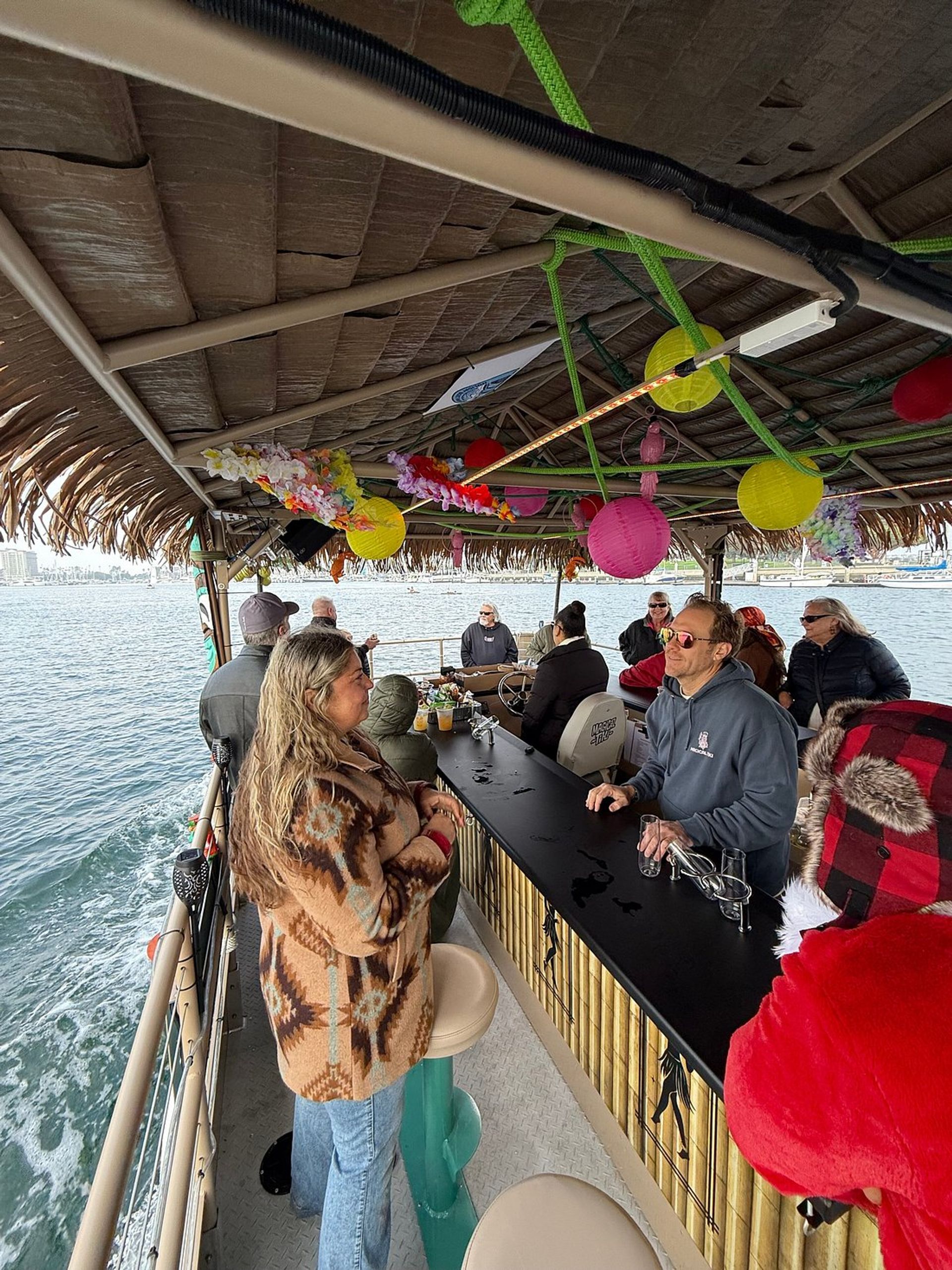 Passengers chatting at a tiki-style floating bar cruise in a coastal harbor — thatched roof decorated with colorful paper lanterns and leis, bamboo counter and ocean views.