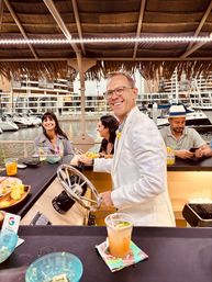 Smiling man in a white jacket steers a floating tiki bar under a thatched roof at a waterfront marina while friends laugh and sip cocktails, with yachts and marina buildings in the background.