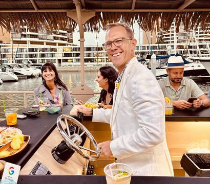 Smiling man in a white jacket steers a floating tiki bar under a thatched roof at a waterfront marina while friends laugh and sip cocktails, with yachts and marina buildings in the background.