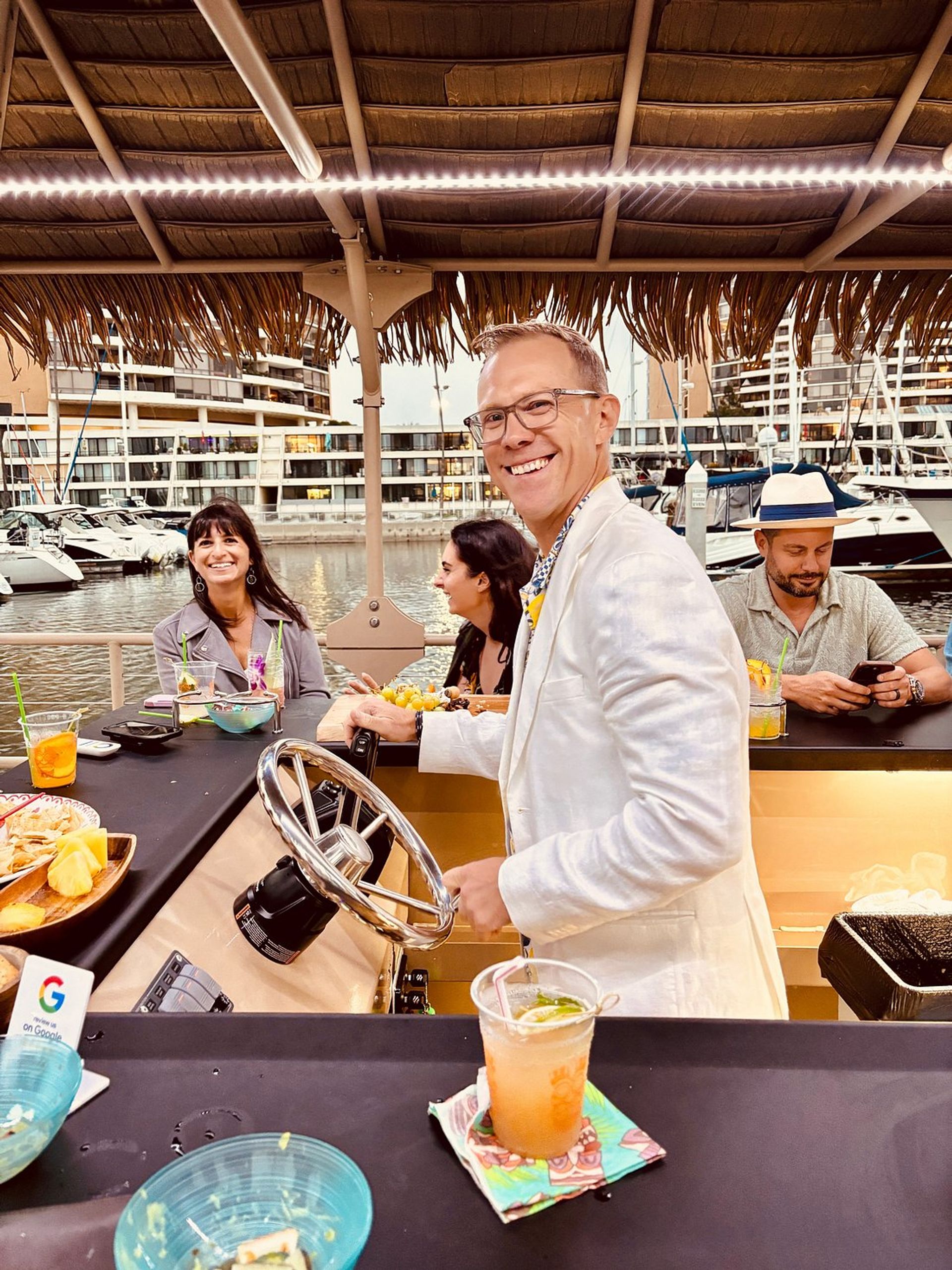 Smiling man in a white jacket steers a floating tiki bar under a thatched roof at a waterfront marina while friends laugh and sip cocktails, with yachts and marina buildings in the background.