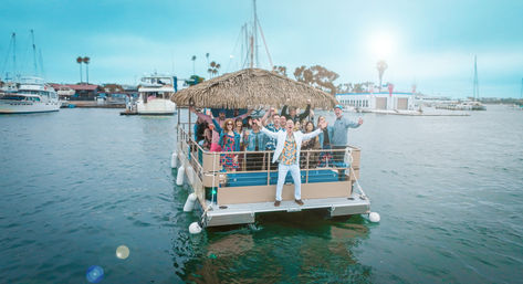 Tiki-style pontoon party boat full of cheering people cruising near a coastal marina with yachts and palm trees