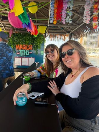 Two smiling friends in sunglasses at a colorful waterfront tiki bar by a marina, under paper lanterns and leis, one holding a blue canned cocktail on the bar.