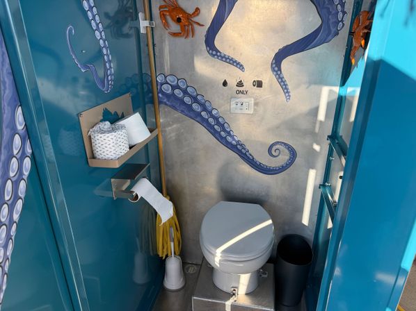 Blue ocean-themed portable toilet interior with painted octopus tentacles and decorative crabs, white toilet, toilet paper shelf and small trash bin, perfect for outdoor events.