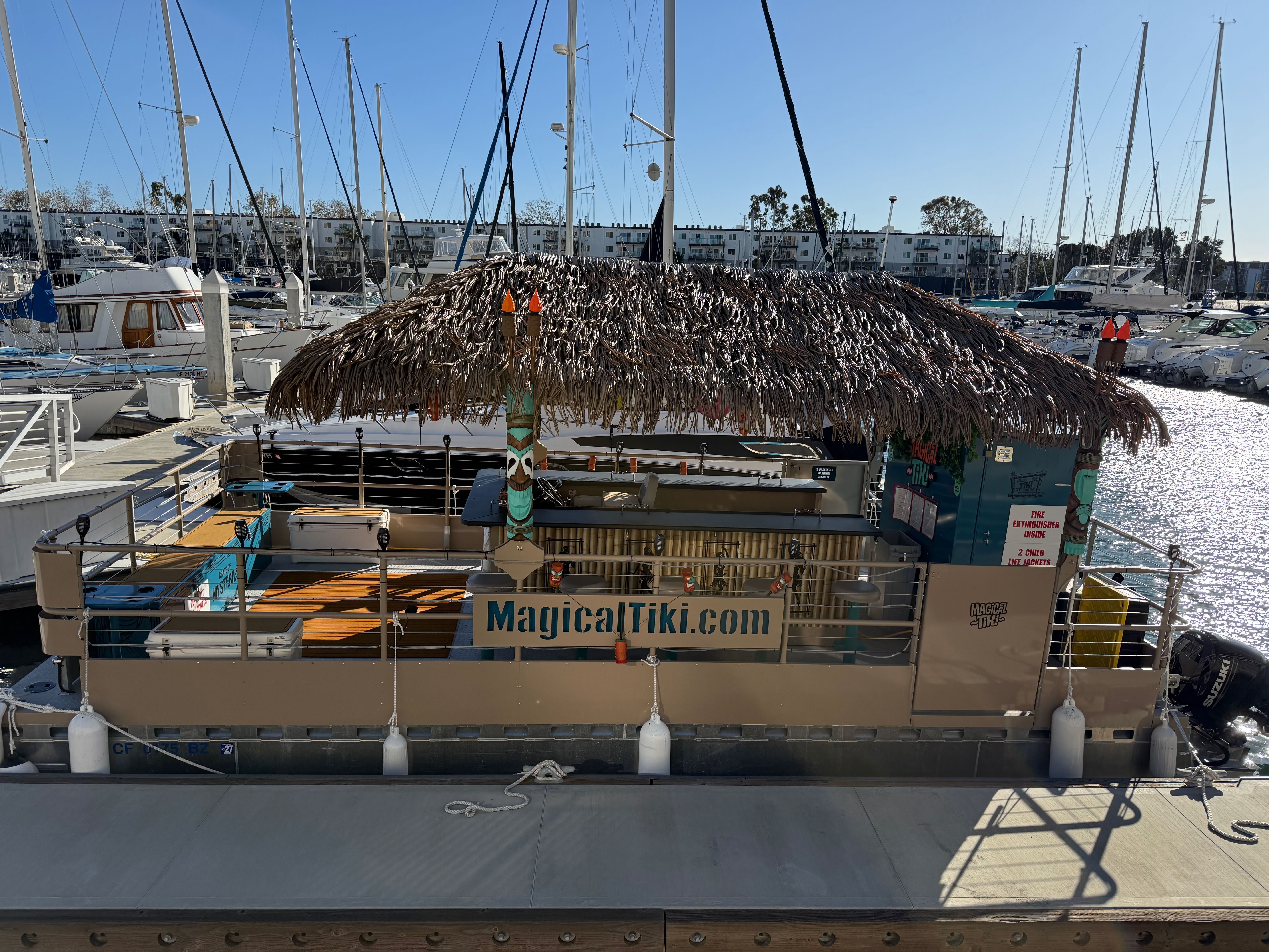 Thatched-roof tiki-bar pontoon docked at a sunny marina, surrounded by sailboats, masts, and sparkling harbor water.