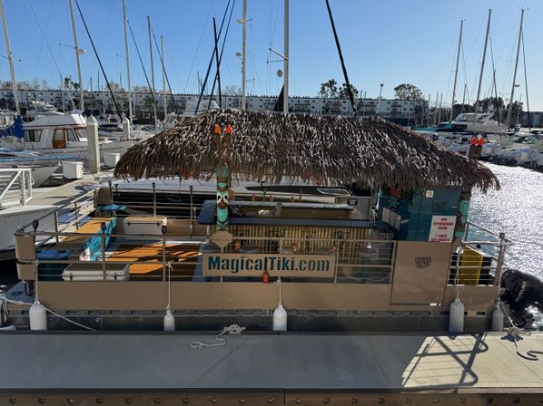 Thatched-roof tiki-bar pontoon docked at a sunny marina, surrounded by sailboats, masts, and sparkling harbor water.