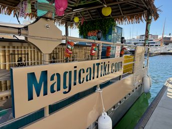 Tiki-themed boat moored at a marina featuring a thatched roof, bamboo panels, colorful tiki masks and paper lanterns hanging from the rail, dock walkway and yachts visible in the background.