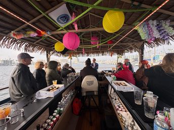 Tiki-style boat bar cruising a marina, passengers seated along a center bar under colorful paper lanterns and leis with a waterfront harbor and moored boats visible outside.