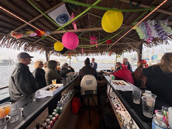 Tiki-style boat bar cruising a marina, passengers seated along a center bar under colorful paper lanterns and leis with a waterfront harbor and moored boats visible outside.