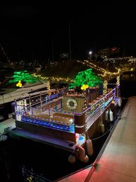 Festive floating tiki bar with a thatched roof, colorful LED rope lights and glowing artificial palm trees, docked at a marina at night.