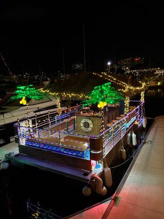 Festive floating tiki bar with a thatched roof, colorful LED rope lights and glowing artificial palm trees, docked at a marina at night.
