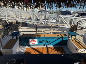 Shaded tiki-roofed boat deck at a marina dock, featuring a bright blue 'Crate of Mysteries' storage chest labeled '20 adult life jackets inside', cushioned benches, cup holders, and docked boats beyond the gangway.
